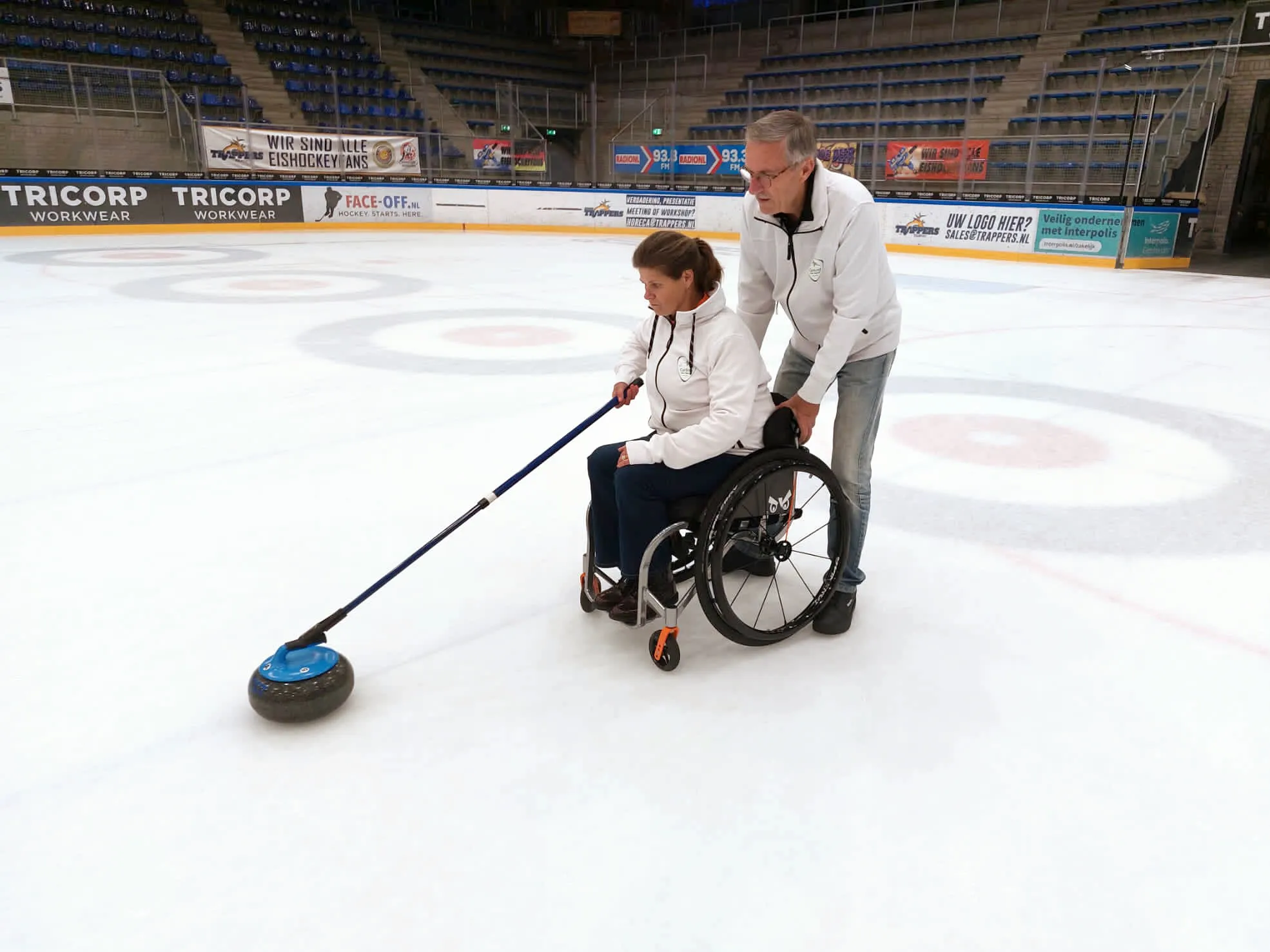 Wheelchair curler delivering a stone with a stick, with a helper behind