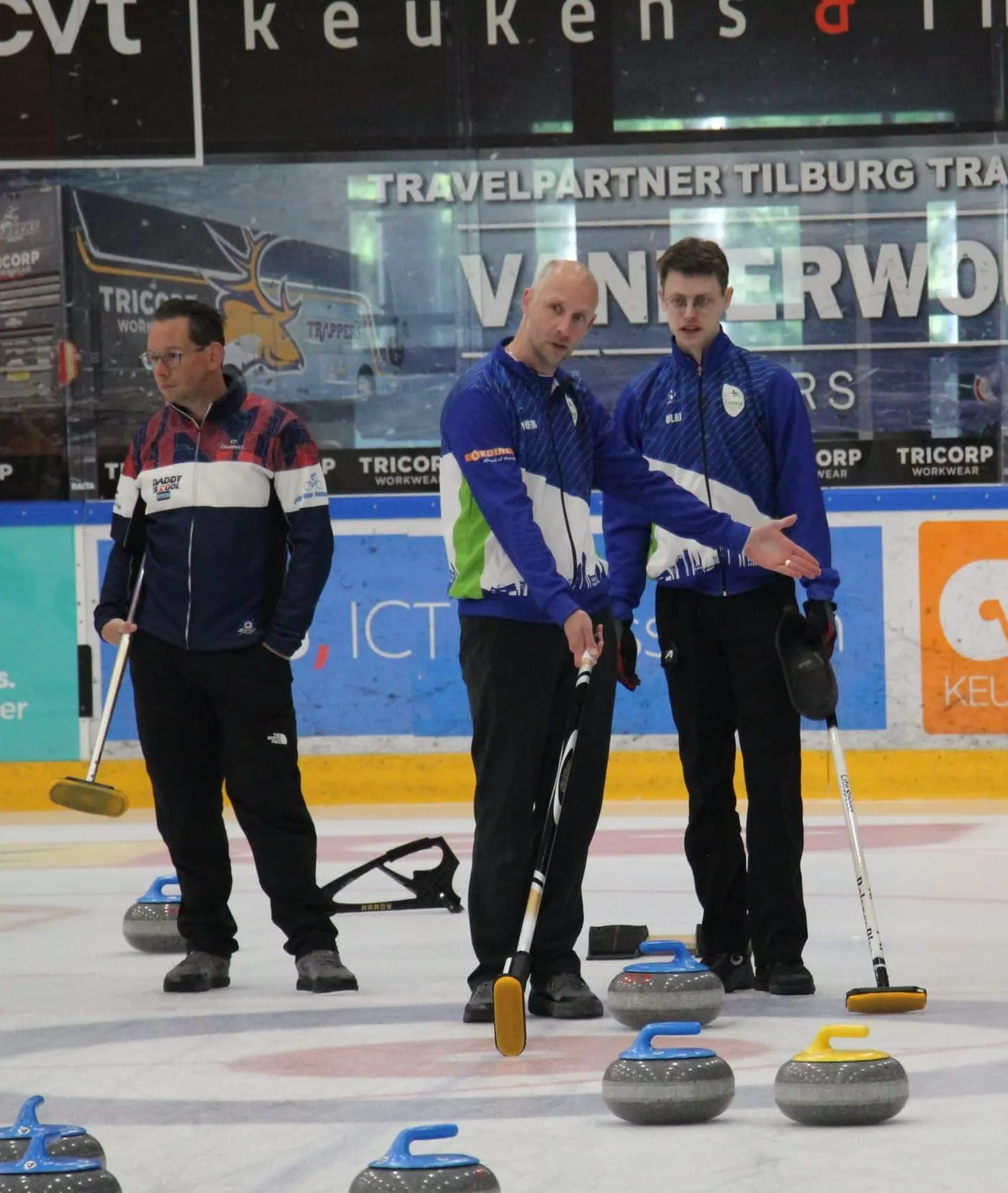 Three curlers in club jackets on the ice next to a row of curling stones